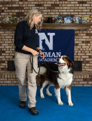 Trainer with a tricolor dog in front of the Neuman K-9 Academy banner